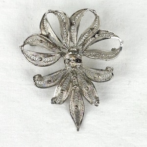 Könnte beinhalten: Silberne Filigranbrosche in floralem Design. Die Brosche hat ein zentrales rundes Element mit strahlenförmigen, durchbrochenen Blütenblättern und gebogenen, bogenartigen Elementen. Die Metallarbeiten sind mit kleinen, dekorativen Mustern versehen.