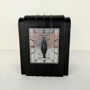 以下が含まれることがあります： シルバーの文字盤が付いた、ヴィンテージの黒いMinute Minderタイマー。タイマーは、分を示すダイヤルを備えたレトロなデザインで、「MINUTE MINDER」の文字が見えます。タイマーはおそらく20世紀半ばのものです。