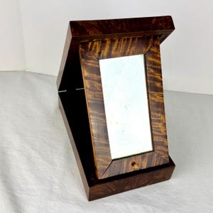 May include: A wooden jewelry box with a hinged lid and a small mirror. The box is made of dark brown wood with a grain pattern. The mirror is rectangular and set within a wooden frame. The box is open, revealing a black interior.