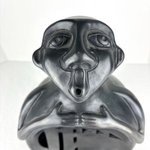 Op de afbeelding: Een glad, zwart keramisch beeld van een gestileerde mensachtige figuur met een ronde kop, grote amandelvormige ogen en een kleine, cirkelvormige mondopening. De figuur heeft de handen bij de borst en een hol, afgerond lichaamsontwerp.
