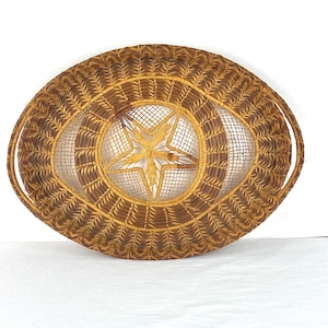 May include: An oval-shaped woven basket featuring a dark brown and golden-tan color palette. The center of the basket displays a prominent five-pointed star design created with intricate open-weave patterns, surrounded by a dense, textured border.