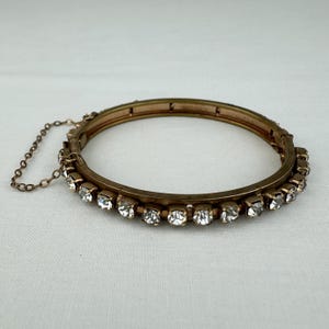 May include: A vintage gold-toned hinged bangle bracelet. The bracelet is adorned with a row of clear, sparkling rhinestones. A delicate chain secures the bracelet's clasp. The bracelet is set against a neutral background.
