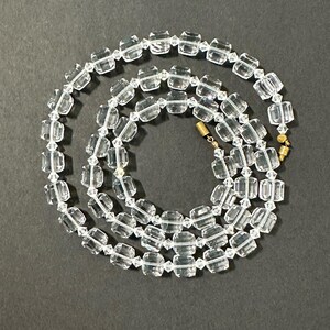 May include: A clear crystal necklace with rectangular beads strung together. The necklace is laid out on a dark grey surface. The clasp is gold-coloured. The beads are faceted, catching the light and creating a sparkling effect.