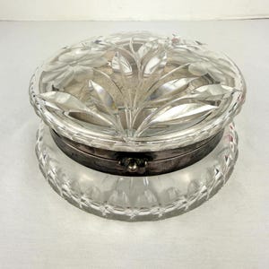 May include: A clear, round, crystal trinket box with a silver-colored metal band. The lid features a floral and leaf design. The base has a decorative, cut-glass pattern. The box is likely used for storing small jewelry items.