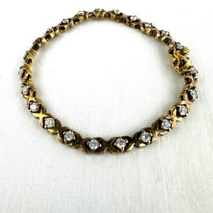 May include: A gold-toned necklace with a repeating pattern of diamond-shaped settings, each holding a clear, round gemstone. The necklace is on a white background, showcasing the jewelry's intricate design and sparkle. The clasp is ornate.