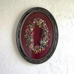 May include: An oval framed floral wreath with a dark red background. The wreath is composed of various colorful flowers and greenery. The frame is dark gray with ornate detailing, adding a vintage aesthetic. The artwork is displayed against a textured white wall.