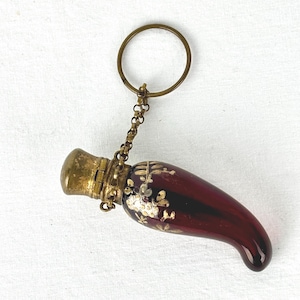 May include: An antique perfume bottle with a deep red body and gold detailing. The bottle has a curved shape and is decorated with gold floral patterns. It features a gold-coloured metal cap and a chain attached to a ring.