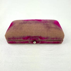 Op de afbeelding: Een rechthoekige, vintage sieradendoos met een bruine stoffen buitenkant en een fuchsia fluwelen bovenkant. De doos is beveiligd met een kleine, zilverkleurige sluiting. De doos is gesloten en staat op een wit oppervlak.
