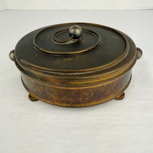 Pode incluir: Uma caixa decorativa redonda em estilo antigo com acabamento em bronze. A caixa tem uma tampa com um design em espiral e uma pequena pega esférica prateada. A caixa tem pequenos pés e duas pegas laterais.