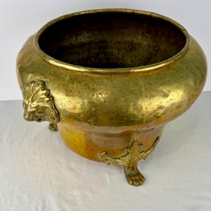 Può includere: Una grande ciotola rotonda in ottone martellato con un'ampia apertura. L'esterno presenta una calda finitura oro metallizzato con patina visibile. È sostenuta da tre piedi ornamentali in ottone fuso a forma di zampe di leone.