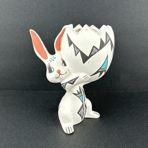 May include: A white ceramic planter shaped like a rabbit holding a large cracked egg. The rabbit features orange inner ears, black painted eyes, and a small floral detail on its forehead. The egg has black geometric patterns and teal accents.