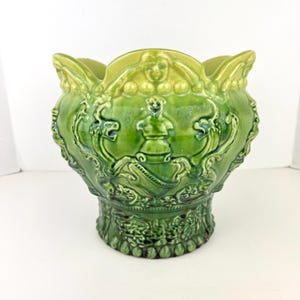 May include: A glossy green ceramic planter featuring a scalloped rim and intricate relief carvings of mythical figures and scrollwork. The vessel has a wide, flared opening that tapers down to a circular base, with a vibrant, multi-tonal finish.