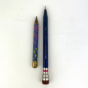 May include: Two pencils on a white background. One is a short, blue pencil with a gold ferrule and a pattern of yellow and pink shapes. The other is a long, dark blue pencil with the text "EAGLE AUTOMATIC PAT. PEND."