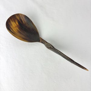 May include: A polished horn spoon featuring a wide, oval-shaped bowl with natural brown and black marbled patterns. The handle is slender and tapers to a point, displaying intricate carved textures and a detailed animal head motif at the neck.