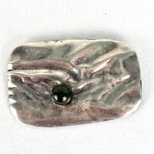 Könnte beinhalten: Rechteckige Brosche aus silberfarbenem Metall mit strukturierter Oberfläche und einem dunkelgrünen, runden Stein in der Mitte. Die Brosche hat ein leicht gehämmertes Aussehen, was ihr einen einzigartigen, handgefertigten Look verleiht.