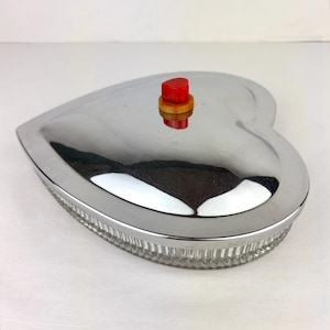 May include: A heart-shaped serving dish with a shiny, silver lid and a clear, textured glass base. The lid has a small, red knob. The dish is likely used for serving food or as a decorative piece.