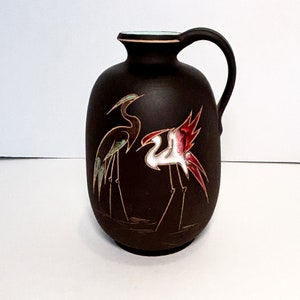 Pode incluir: Um jarro de cerâmica castanho-escuro com acabamento mate e uma única pega. O corpo está decorado com duas ilustrações estilizadas de grous, uma em tons de azul-petróleo e castanho, e a outra em branco, vermelho e castanho, na água.