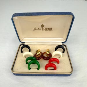 May include: A vintage jewelry set by Trifari, presented in a blue box. The set includes gold-toned hoop earrings and various colored clip-on earrings in black, white, green, and red. The box is lined with a cream-colored fabric.