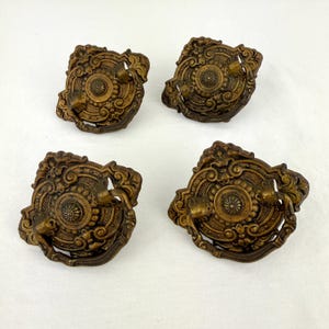 May include: Four antique bronze drawer pulls with ornate, detailed designs. Each pull features a central circular element with radiating patterns, surrounded by scrolling embellishments and a curved handle. The pulls are arranged on a white surface.