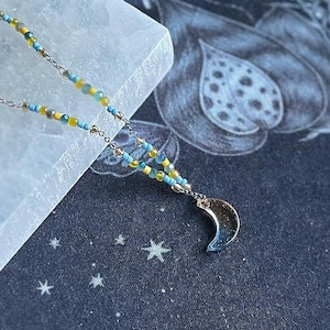 Puede incluir: Un collar de cadena plateada con cuentas azules y amarillas y un dije de media luna.