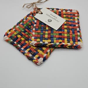 May include: Two square, woven hot pads in a colorful pattern of orange, yellow, blue, green, and brown. A tag attached with twine reads "handcrafted by DDRocksPhxAZ". The pads are made in the USA.