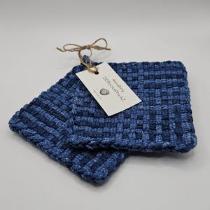 May include: Two blue woven potholders with a tag attached by twine. The tag reads "handcrafted by DDRocksPhxAZ". The potholders are made of a woven material in shades of blue.