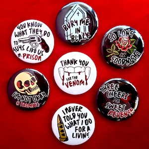 May include: Seven circular pin-back buttons featuring various graphic designs and text. Designs include a revolver, a coffin, a rose, vampire fangs, a skull, and a bullet. Text includes phrases like Bury Me In Black, Thank You For The Venom, and I'm Not Okay.