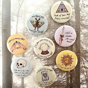 May include: Set of ten 1-inch button pins with various illustrations and text. The pins feature a skull, a butterfly, a sun, a crystal ball, a hand with a sword, a hand with a triangle, and a tarot card. The text on the pins includes phrases like "I'll show you every version of yourself tonight", "Take me to the lakes where all the poets want to die", and "What a shame she went mad".