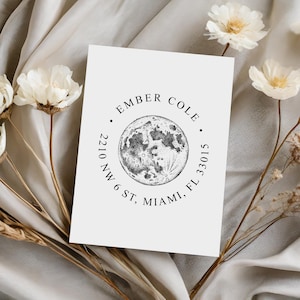 May include: A white card featuring a black and white illustration of the moon, with the text "EMBER COLE" and address details. The card is surrounded by white flowers and dried plants, set against a neutral fabric backdrop.