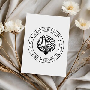 May include: A white card featuring a circular stamp design with the name "Adeline Reese" and address details. The stamp includes a detailed seashell illustration in the center, surrounded by text. The card is set against a neutral, textured background with floral accents.