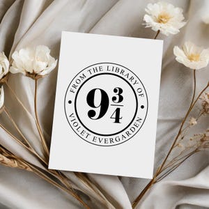 May include: A white card with a black circular design featuring the text "FROM THE LIBRARY OF VIOLET EVERGARDEN" surrounding the number "9 3/4". The card is decorated with white flowers and dried stems.