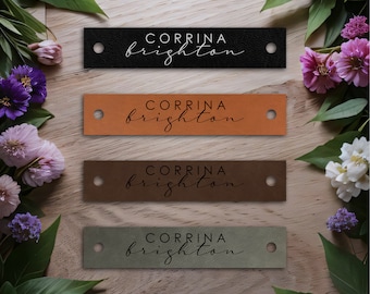 Op de afbeelding: Vier leren labels met de naam "Corrina Brighton" in een cursief schrift. De labels zijn in zwart, bruin, oranje en groen.