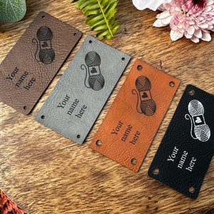 May include: Four leather labels with a yarn ball and heart design. The labels are in brown, gray, orange, and black. Each label has the text "Your name here" printed on it.