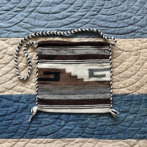 May include: A square woven shoulder bag featuring a geometric pattern in shades of brown, cream, black, and gray. The bag has a braided black and white strap and matching braided trim along the edges with small tassels at the bottom corners.
