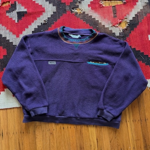 May include: A purple fleece sweatshirt with a crew neck and long sleeves. The collar and chest pocket feature a patterned design with teal, brown, and orange accents. The sweatshirt has a small logo on the chest.