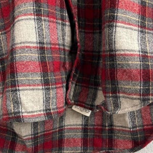 May include: A long-sleeve button-down shirt featuring a plaid pattern in shades of red, charcoal gray, and off-white. The garment includes a front placket with metallic buttons and a small white care label attached to the bottom hem area.
