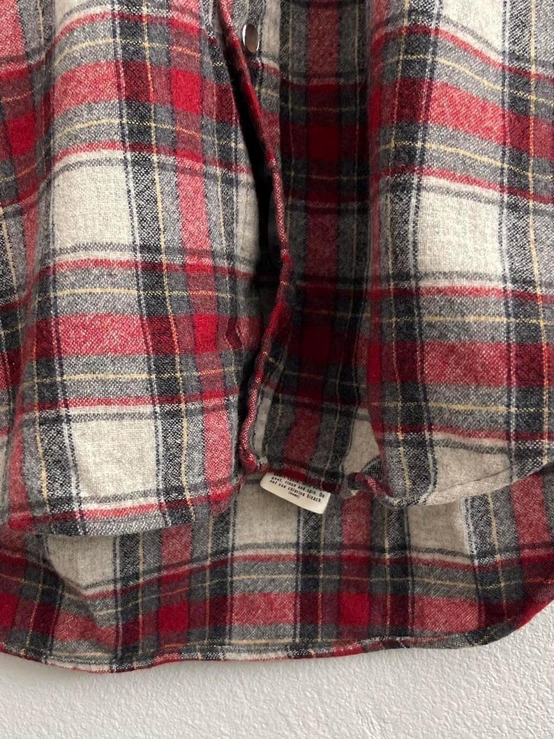 May include: A long-sleeve button-down shirt featuring a plaid pattern in shades of red, charcoal gray, and off-white. The garment includes a front placket with metallic buttons and a small white care label attached to the bottom hem area.