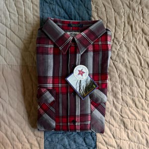 May include: A long-sleeve button-down shirt featuring a red, grey, and black plaid pattern. The shirt includes two chest pockets and a tag that reads Crafted with Pride in U.S.A. and Fieldmaster. The collar is folded and the fabric has a visible texture.