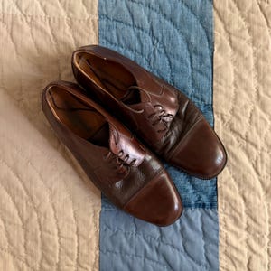 May include: A pair of dark brown leather oxford shoes featuring a cap toe design, a lace-up closure with thin matching laces, and a smooth finish on the toe box with a slightly textured leather panel along the sides and the eyelet area.