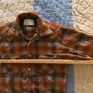 K&ouml;nnte beinhalten: Ein lang&auml;rmeliges Pendleton-Hemd mit orangefarbenem, braunem und grauem Karomuster, Kragen, zwei Brusttaschen und Druckkn&ouml;pfen. Ein Holzlineal liegt quer &uuml;ber der Brust und zeigt eine Breitenmessung von etwa 58 Zentimetern an.