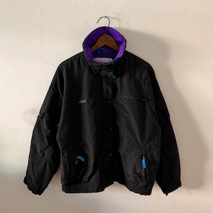 May include: A black Columbia jacket featuring a high collar with a vibrant purple interior lining. The jacket includes a front zipper closure, long sleeves with elastic cuffs, and bright turquoise accents on the zipper pulls and logo.