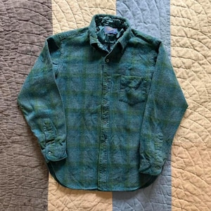 May include: A long-sleeved button-down shirt in a teal and dark green plaid pattern. The shirt features a standard collar, a single chest pocket on the left side, and buttoned cuffs. A blue Pendleton brand label is visible inside the collar.
