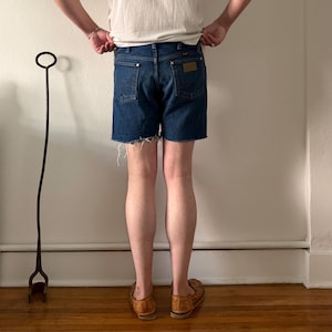 May include: Dark blue denim shorts featuring a raw, frayed hemline, a classic five-pocket design, a leather-style patch on the back right pocket, and copper-toned rivets. The shorts are worn with a plain white t-shirt and brown woven leather shoes.