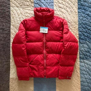 May include: A bright red puffer jacket with a high collar, long sleeves, and a full-length front zipper. The jacket features horizontal quilted sections and a small white tag attached to the zipper pull with the text Timberline and 01/19/97.