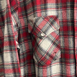 May include: A long-sleeve button-up shirt featuring a red, grey, and black plaid pattern. The shirt includes a single chest pocket with a pointed flap, secured by a silver-toned metal snap button. The fabric displays a soft, brushed texture.