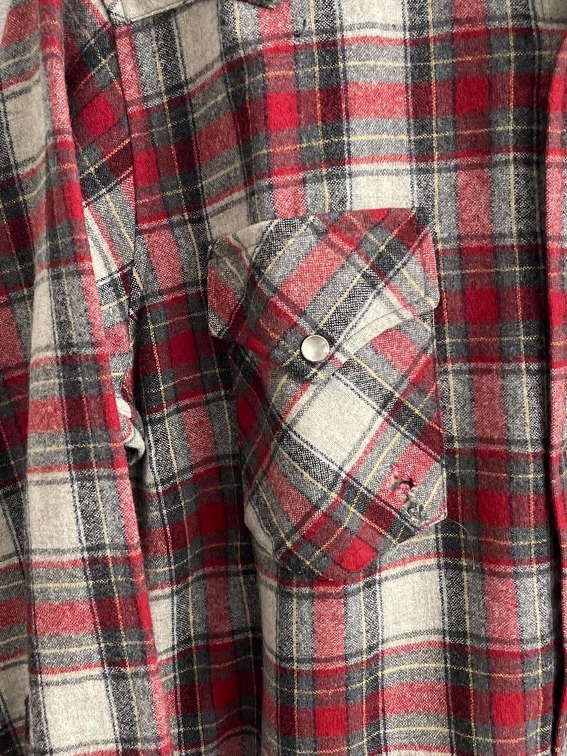 May include: A long-sleeve button-up shirt featuring a red, grey, and black plaid pattern. The shirt includes a single chest pocket with a pointed flap, secured by a silver-toned metal snap button. The fabric displays a soft, brushed texture.