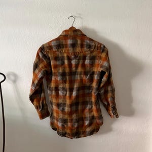 K&ouml;nnte beinhalten: Ein lang&auml;rmeliges Hemd mit Knopfleiste und einem Karomuster in Orange-, Braun- und Graut&ouml;nen. Das Hemd hat einen spitzen Kragen, eine R&uuml;ckenpasse im Western-Stil und gekn&ouml;pfte Manschetten und h&auml;ngt an einem Kleiderb&uuml;gel.