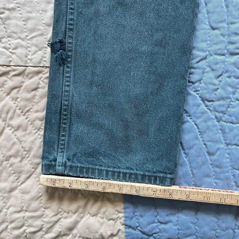 Puede incluir: Una pernera de pantal&oacute;n vaquero de color verde azulado con una peque&ntilde;a zona deshilachada cerca de la costura lateral. Una regla de madera est&aacute; colocada a lo largo del dobladillo inferior, mostrando una anchura de unos 28 cm.