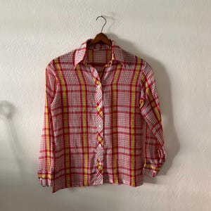 May include: A long-sleeve button-down shirt featuring a plaid pattern in shades of red, yellow, and white. The shirt has a pointed collar, a front button placket with white buttons, and buttoned cuffs on the sleeves, all displayed on a hanger.