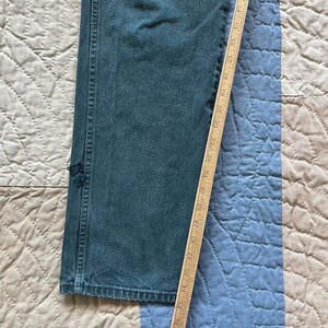 Puede incluir: Unos vaqueros de mezclilla en color verde azulado oscuro con un agujero deshilachado visible en la costura lateral. Una regla de madera est&aacute; colocada verticalmente a lo largo de la pierna, mostrando una medida de 35,5 cm a 83,8 cm.